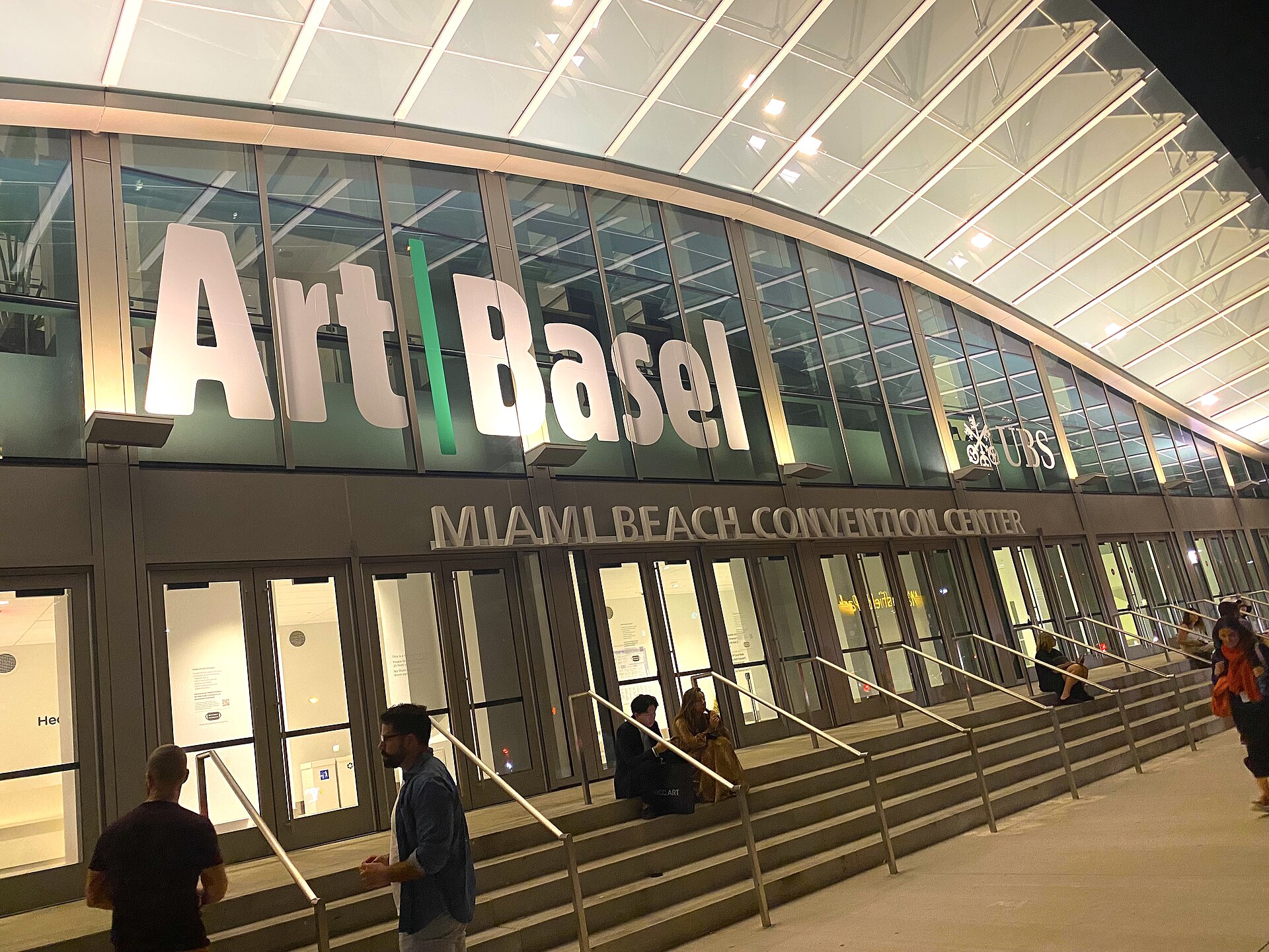 The illuminated Miami Beach Convention Center at night with large Art Basel lettering above the entrance and visitors on the steps