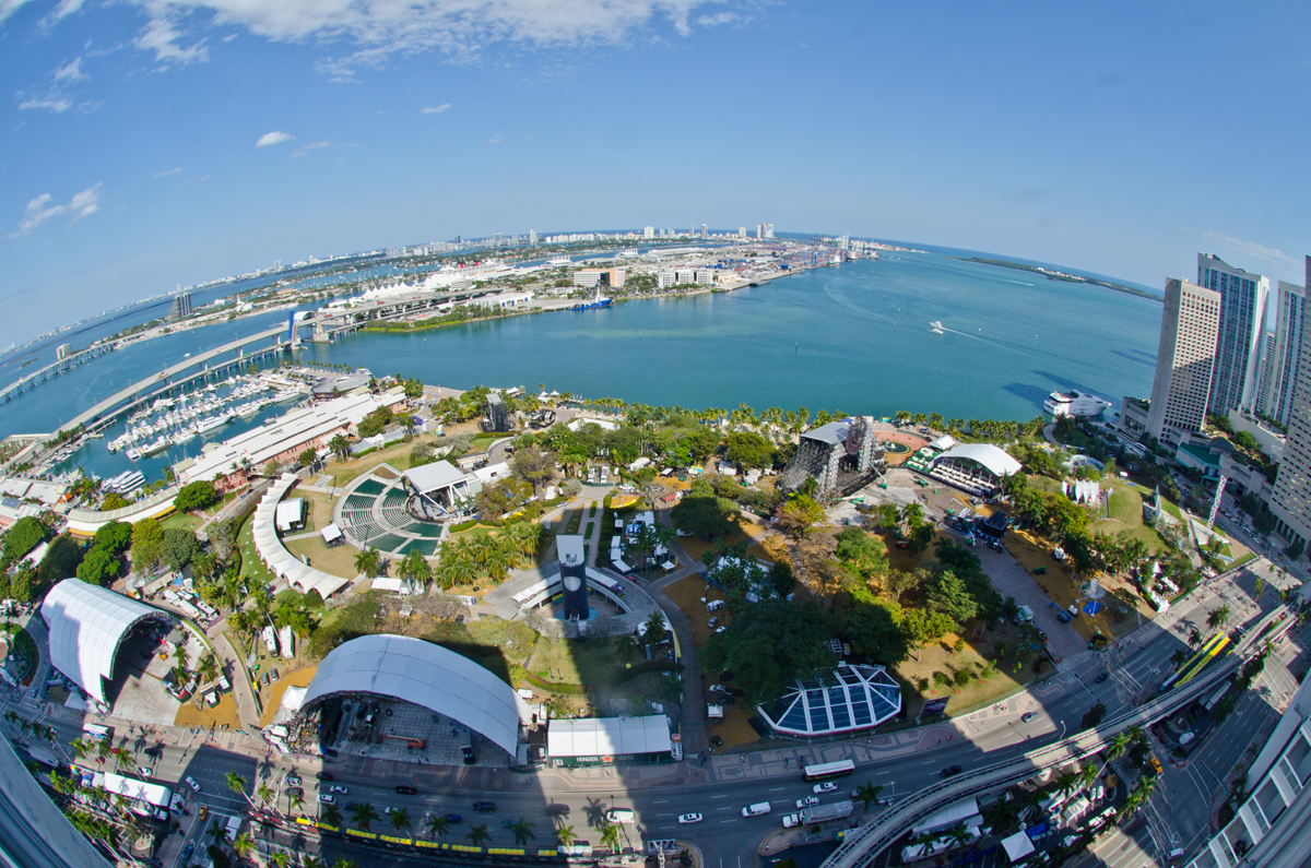 An aerial fisheye view of Bayfront Park set up with stages and tents for Ultra Music Festival, with Biscayne Bay and PortMiami beyond