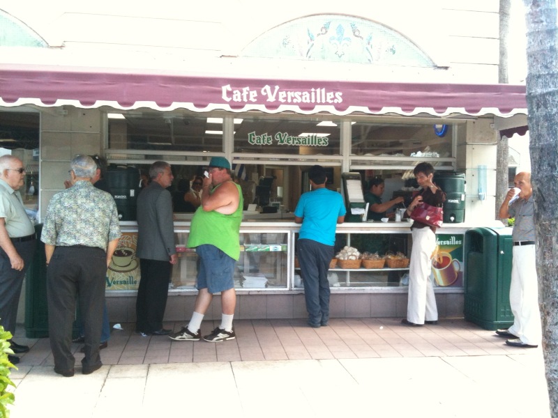 People standing at the outdoor coffee counter of Cafe Versailles in Little Havana, under a maroon awning
