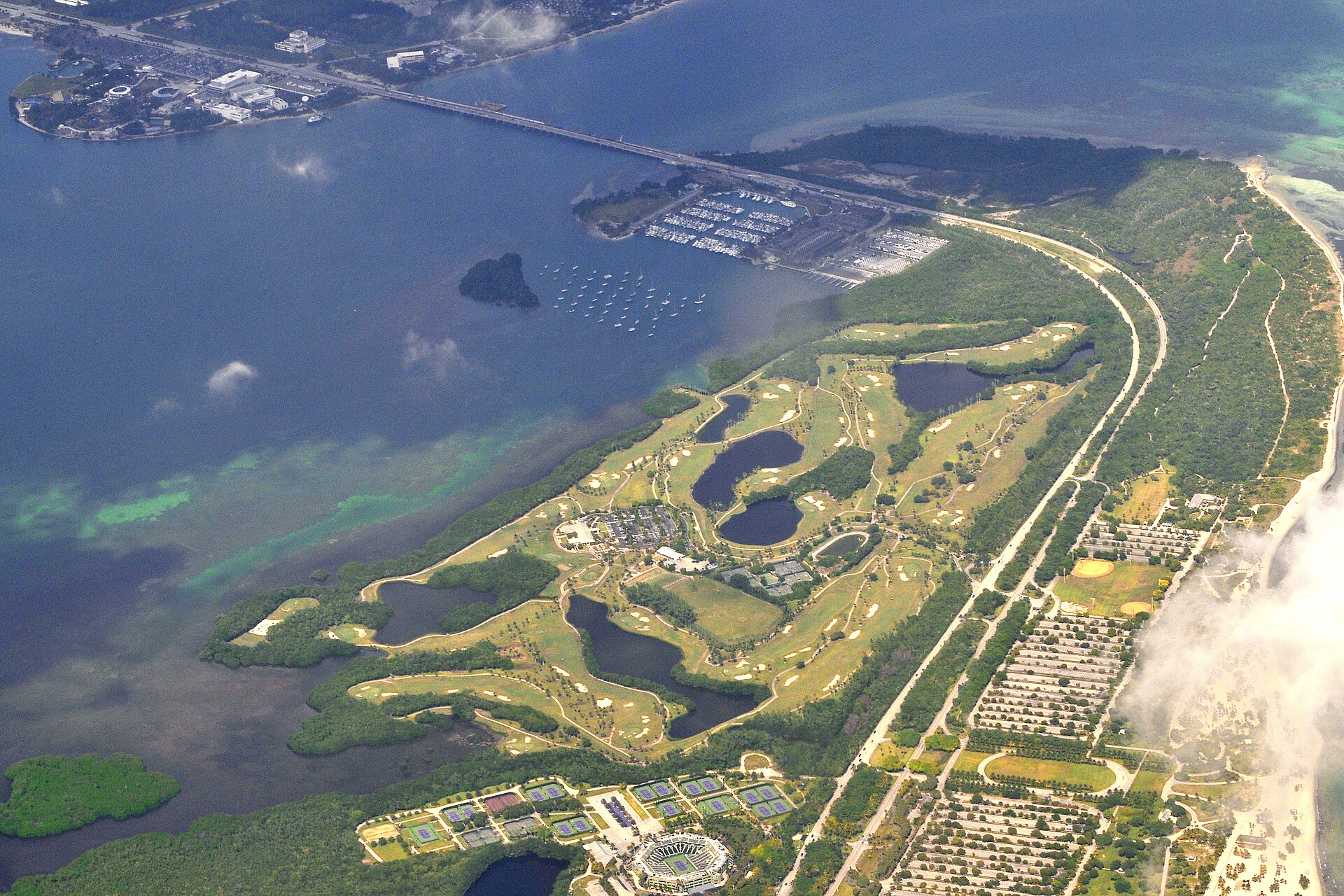 Aerial view of Crandon Park on Key Biscayne, with the golf course's fairways and lakes between the bay and the park road, the old tennis center below and the Rickenbacker Causeway beyond