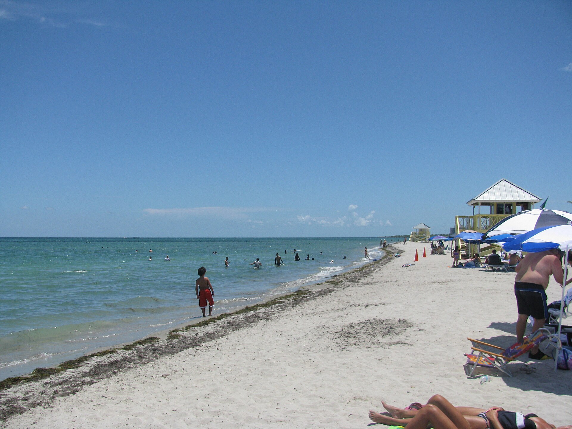 Families wading in shallow calm water at Crandon Park beach on Key Biscayne, with a yellow lifeguard tower and umbrellas along the sand