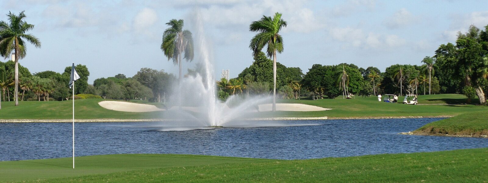 A green and flagstick beside a lake with a fountain, bunkers and royal palms on one of the resort golf courses at Doral