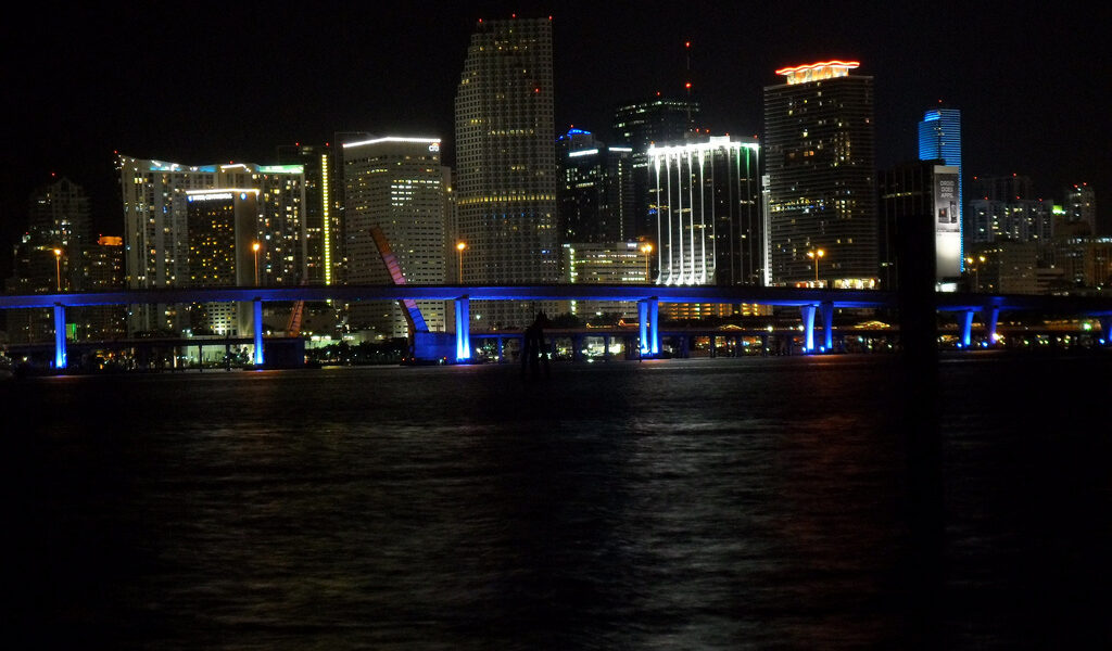 The downtown Miami skyline at night across Biscayne Bay with the illuminated Port Boulevard bridge in the foreground