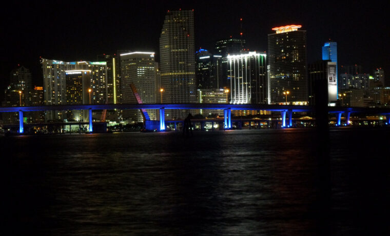 The downtown Miami skyline at night across Biscayne Bay with the illuminated Port Boulevard bridge in the foreground