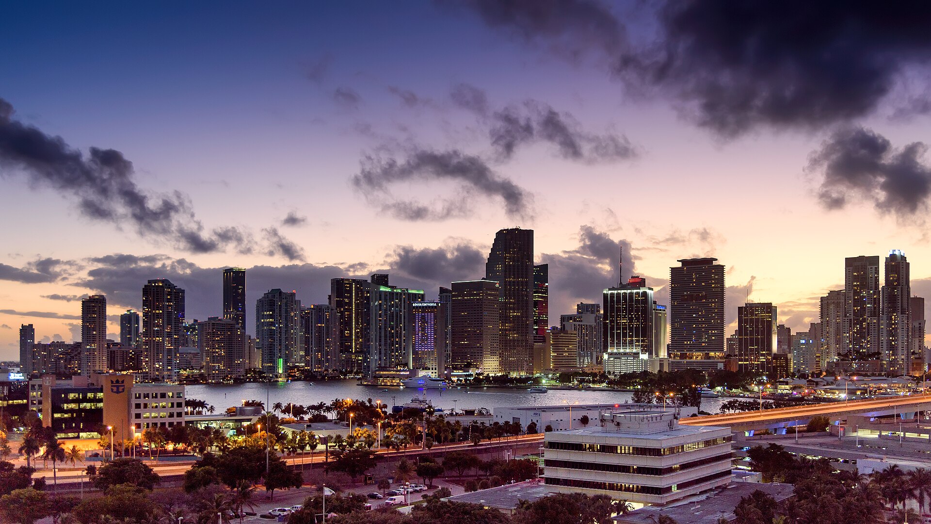 The downtown Miami skyline at dusk seen across the water from Dodge Island, with the port's office buildings and access road in the foreground