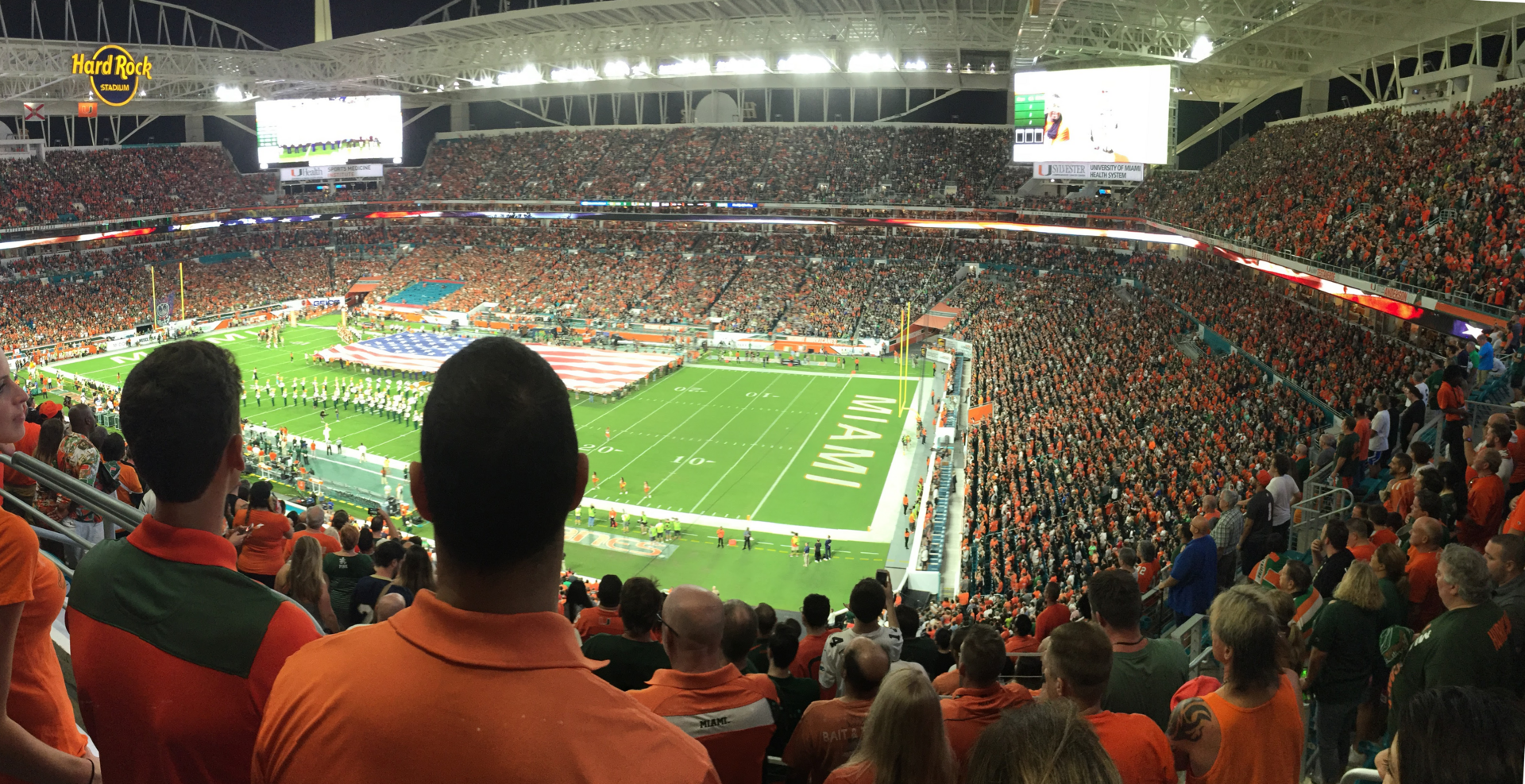 A packed Hard Rock Stadium at night during a Miami Hurricanes game, seen from the upper level, with a giant American flag on the field