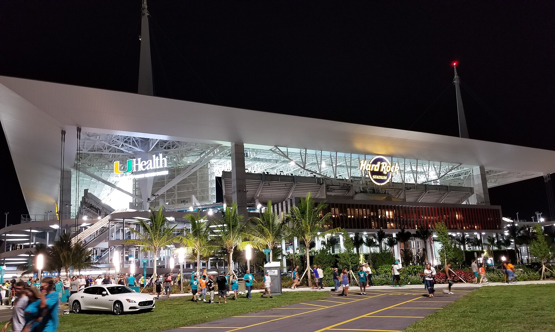 Hard Rock Stadium in Miami Gardens lit up at night, with fans walking past palm trees outside