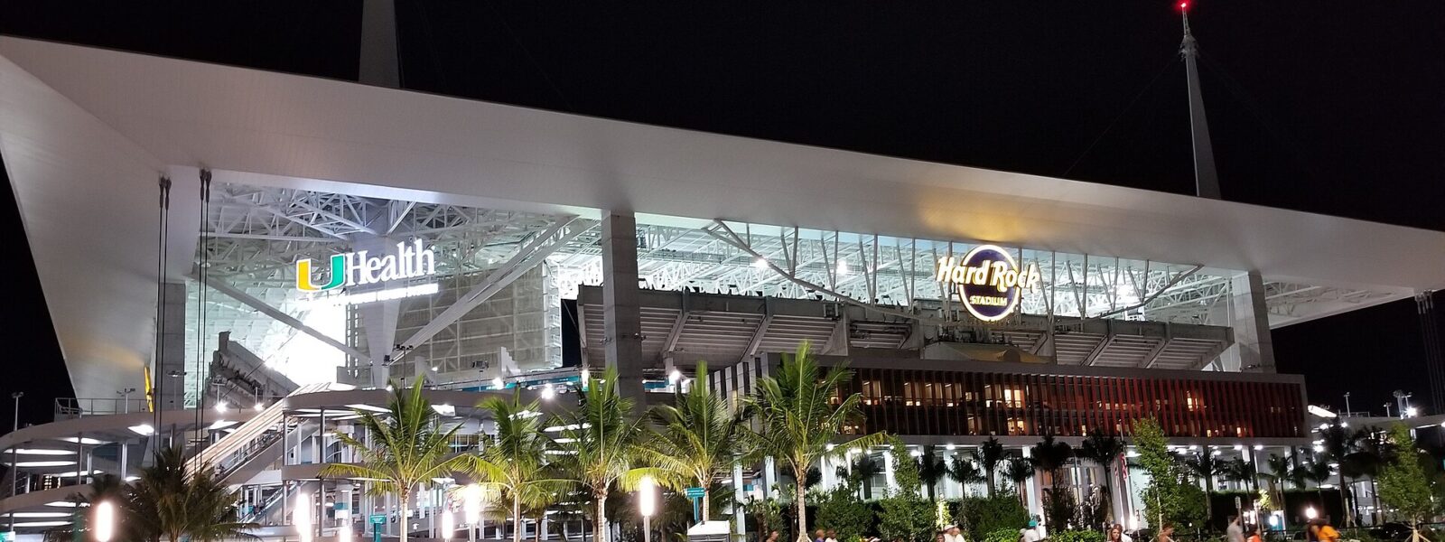 Hard Rock Stadium lit up at night under its white canopy and masts, with Dolphins fans in teal walking out across the lawn