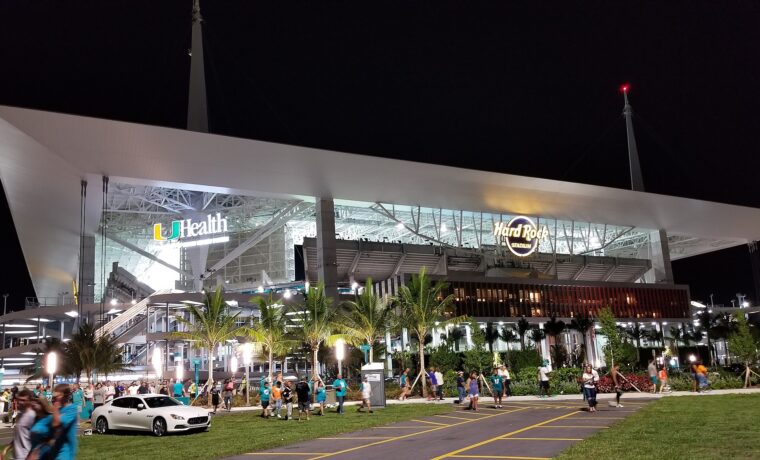 Hard Rock Stadium lit up at night under its white canopy and masts, with Dolphins fans in teal walking out across the lawn