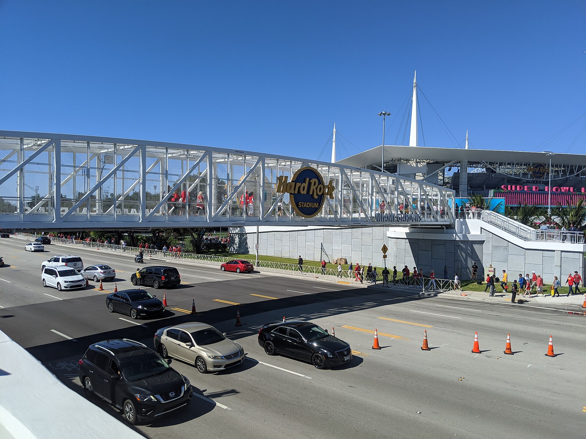 Fans crossing a white pedestrian bridge with a Hard Rock Stadium sign over a busy road toward the stadium on Super Bowl weekend