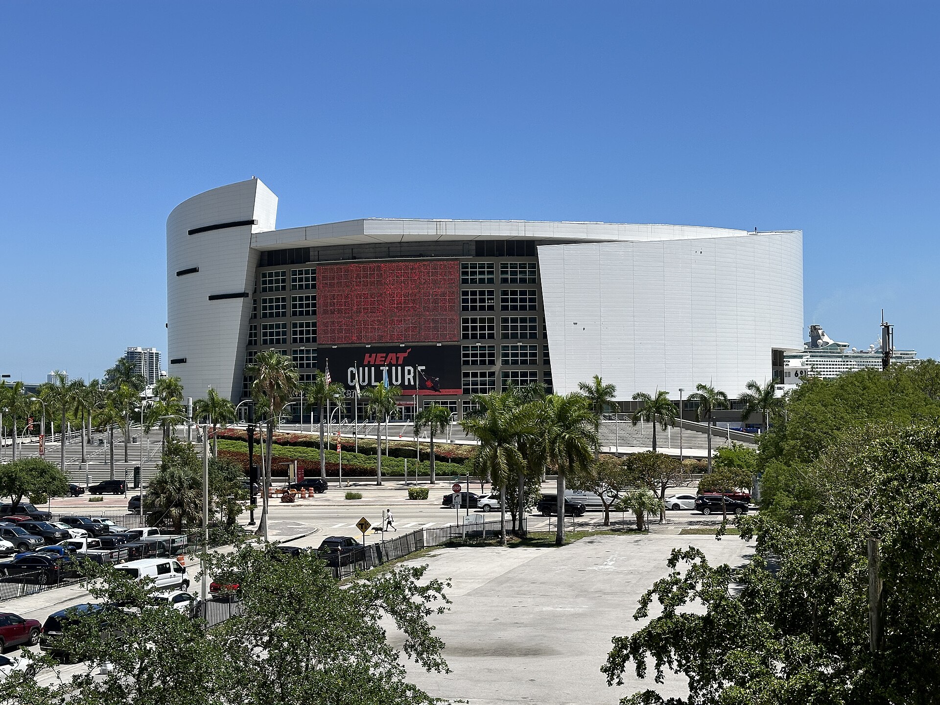 The white curved exterior of Kaseya Center with a Heat Culture banner across the front, palm trees below and a cruise ship docked behind it