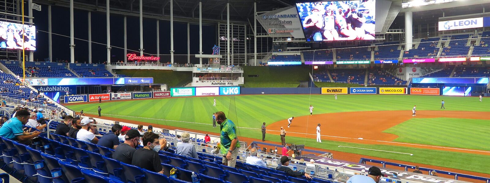 Fans in the blue lower-level seats at loanDepot park during a night game, with the roof closed and the outfield and video boards beyond