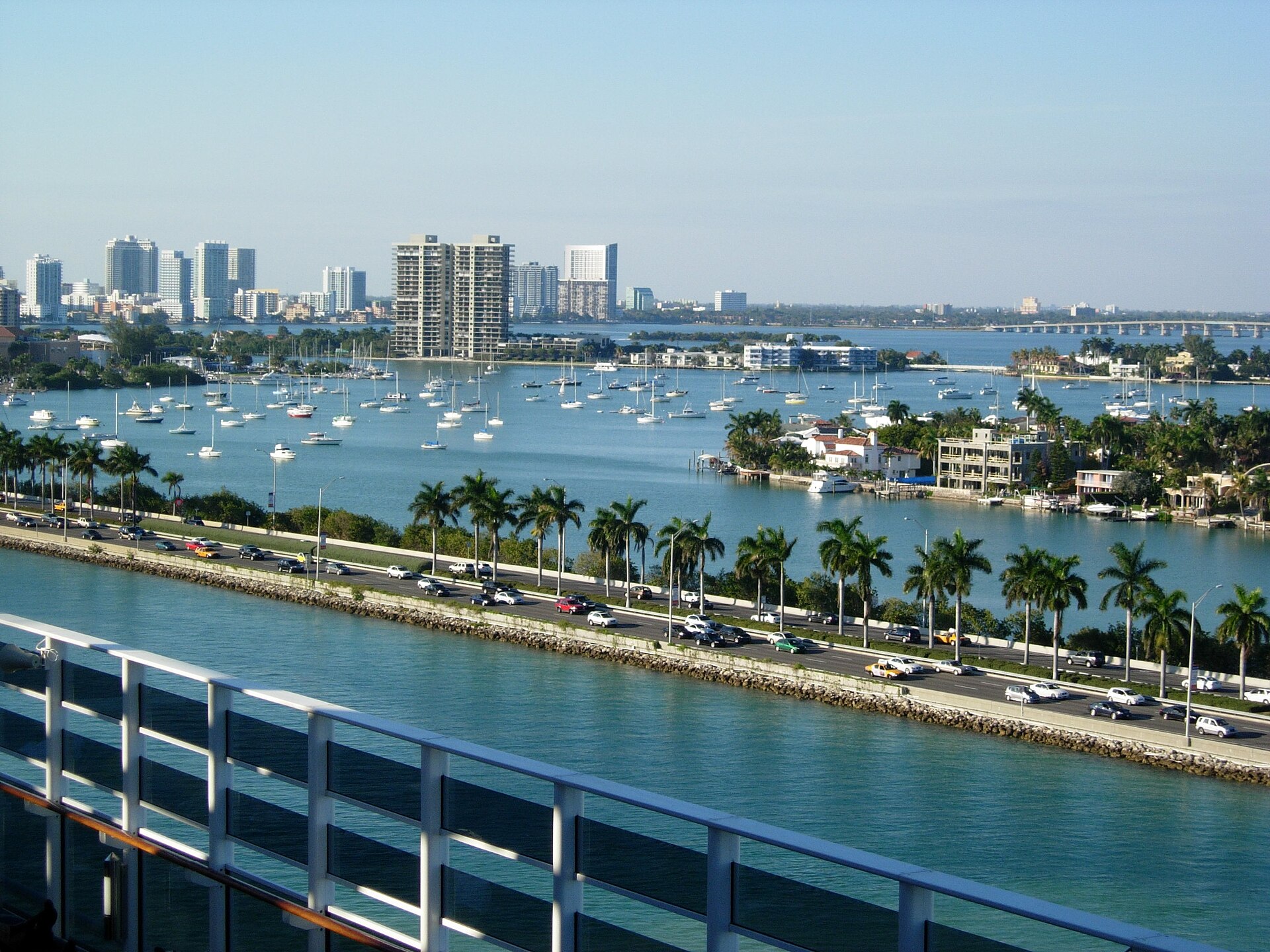 View from the deck of a cruise ship docked at PortMiami, looking across the MacArthur Causeway traffic toward sailboats and Miami Beach
