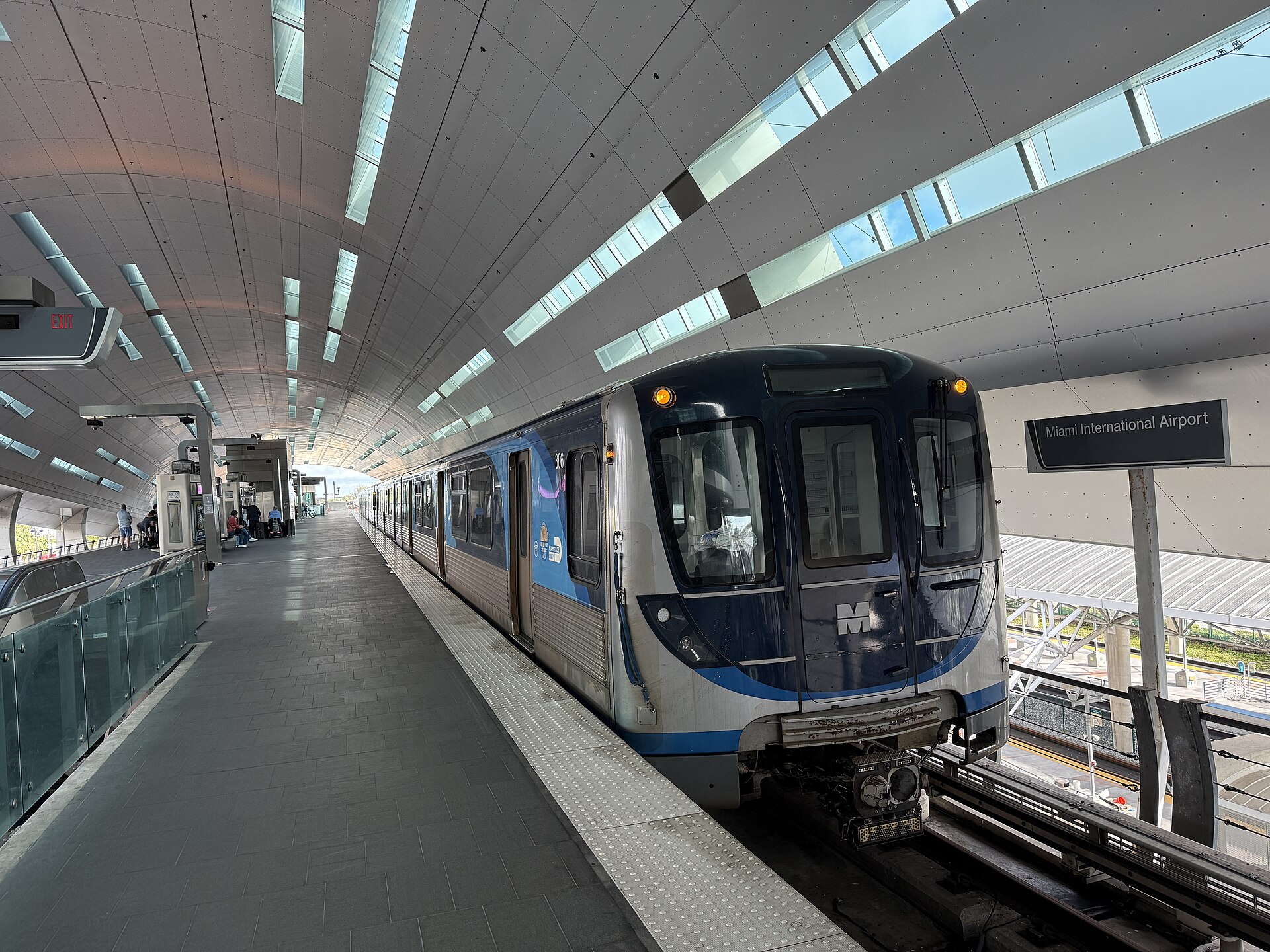A blue and silver Metrorail train waiting at the Miami International Airport station platform under a curved roof with skylights