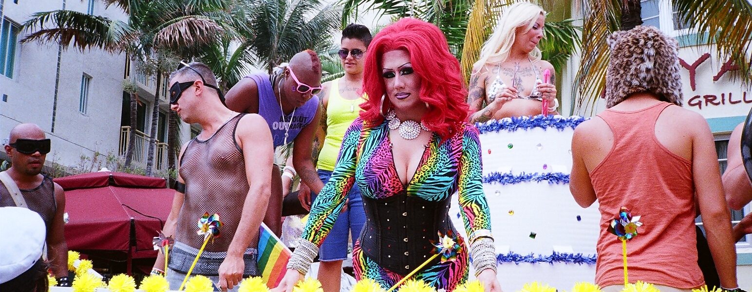 A Miami Beach Pride float with drag performers moving along Ocean Drive past palms and Art Deco buildings