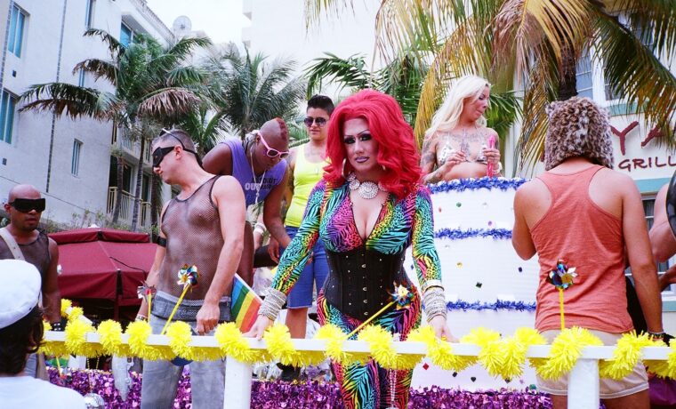 A Miami Beach Pride float with drag performers moving along Ocean Drive past palms and Art Deco buildings