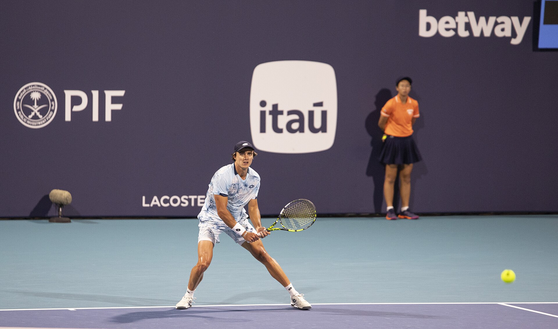 A male player in a light blue patterned shirt waiting to return serve under the lights at the Miami Open, with a ball kid behind him