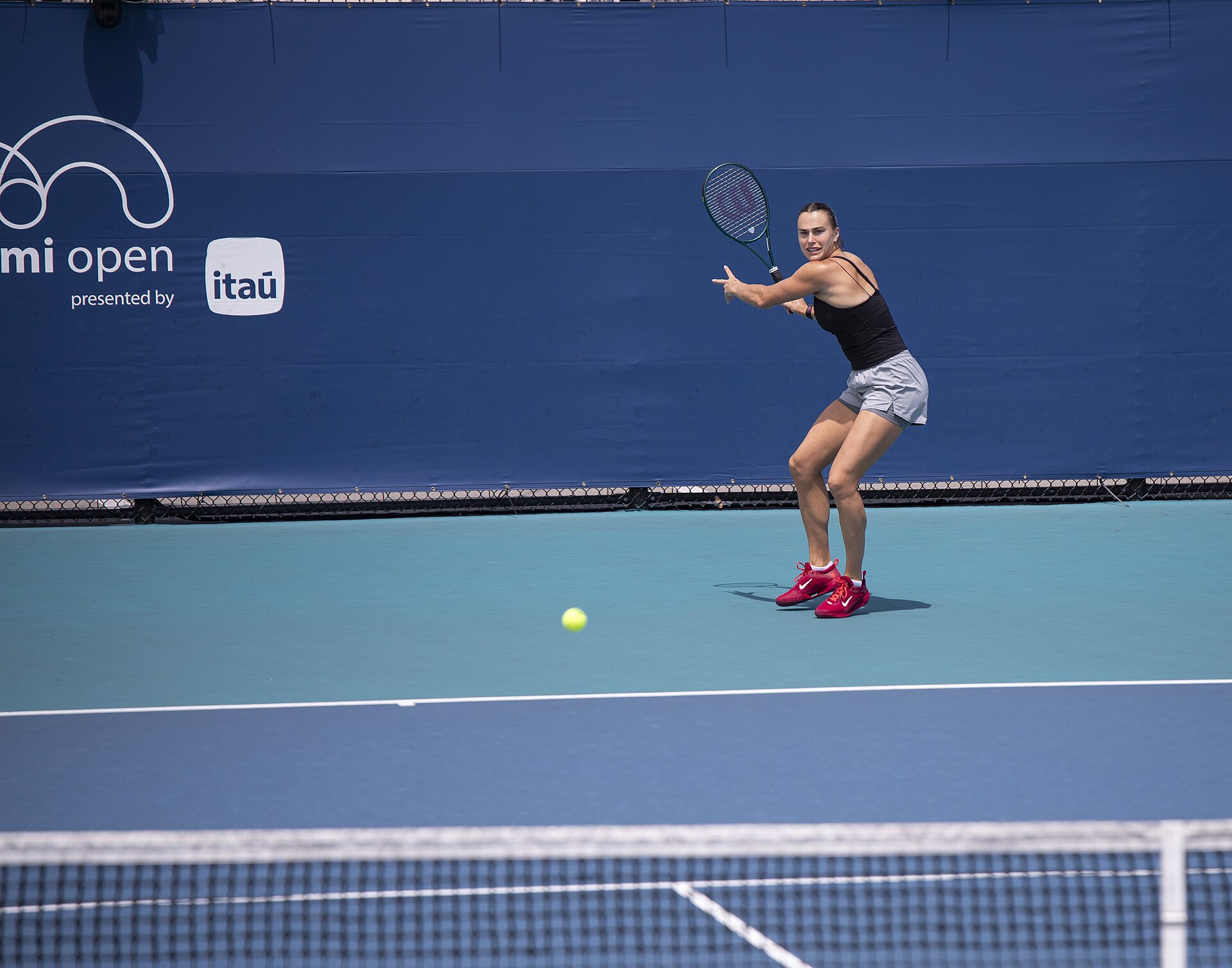 A player hitting a forehand on a blue practice court at the Miami Open, with a dark blue windscreen behind her