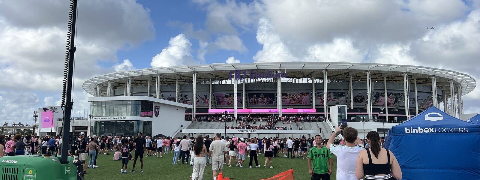 Fans in pink Inter Miami shirts on the lawn outside the South Gate and team store of Nu Stadium under a white canopy roof