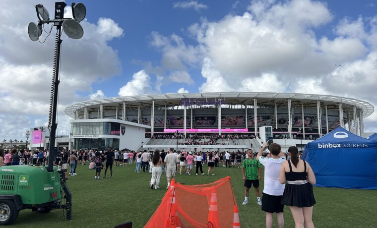 Fans in pink Inter Miami shirts on the lawn outside the South Gate and team store of Nu Stadium under a white canopy roof