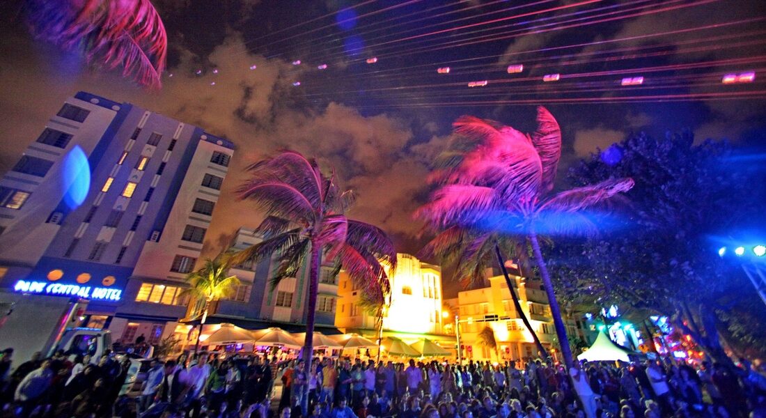 A dense evening crowd on Ocean Drive under palm trees lit pink and blue, with the Park Central Hotel on the left
