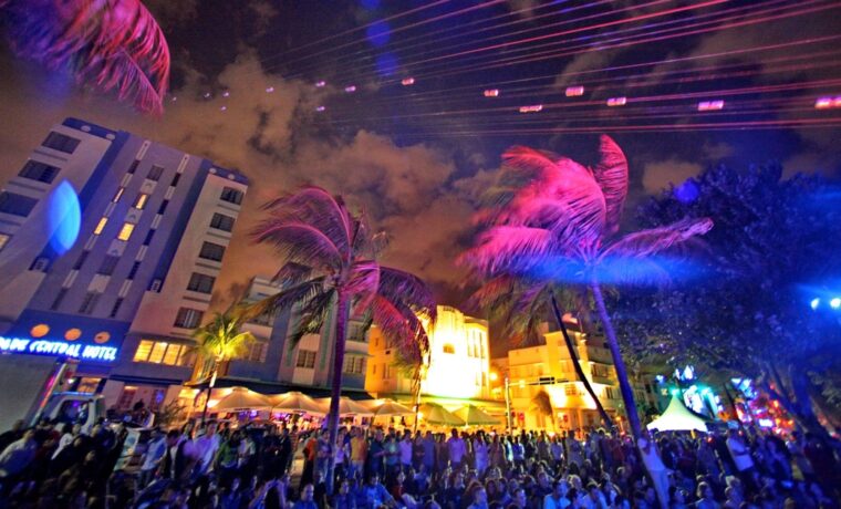 A dense evening crowd on Ocean Drive under palm trees lit pink and blue, with the Park Central Hotel on the left