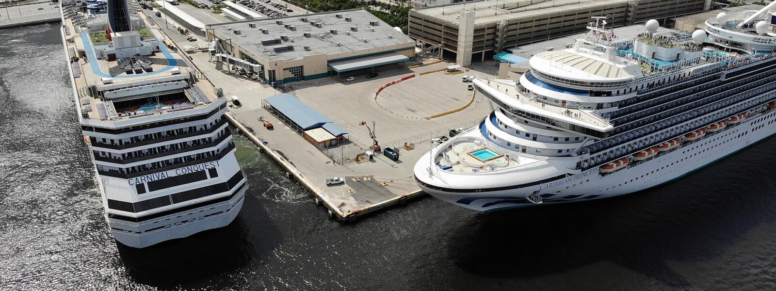 Aerial view of two cruise ships berthed at a Port Everglades terminal, with a parking garage and the Fort Lauderdale skyline behind