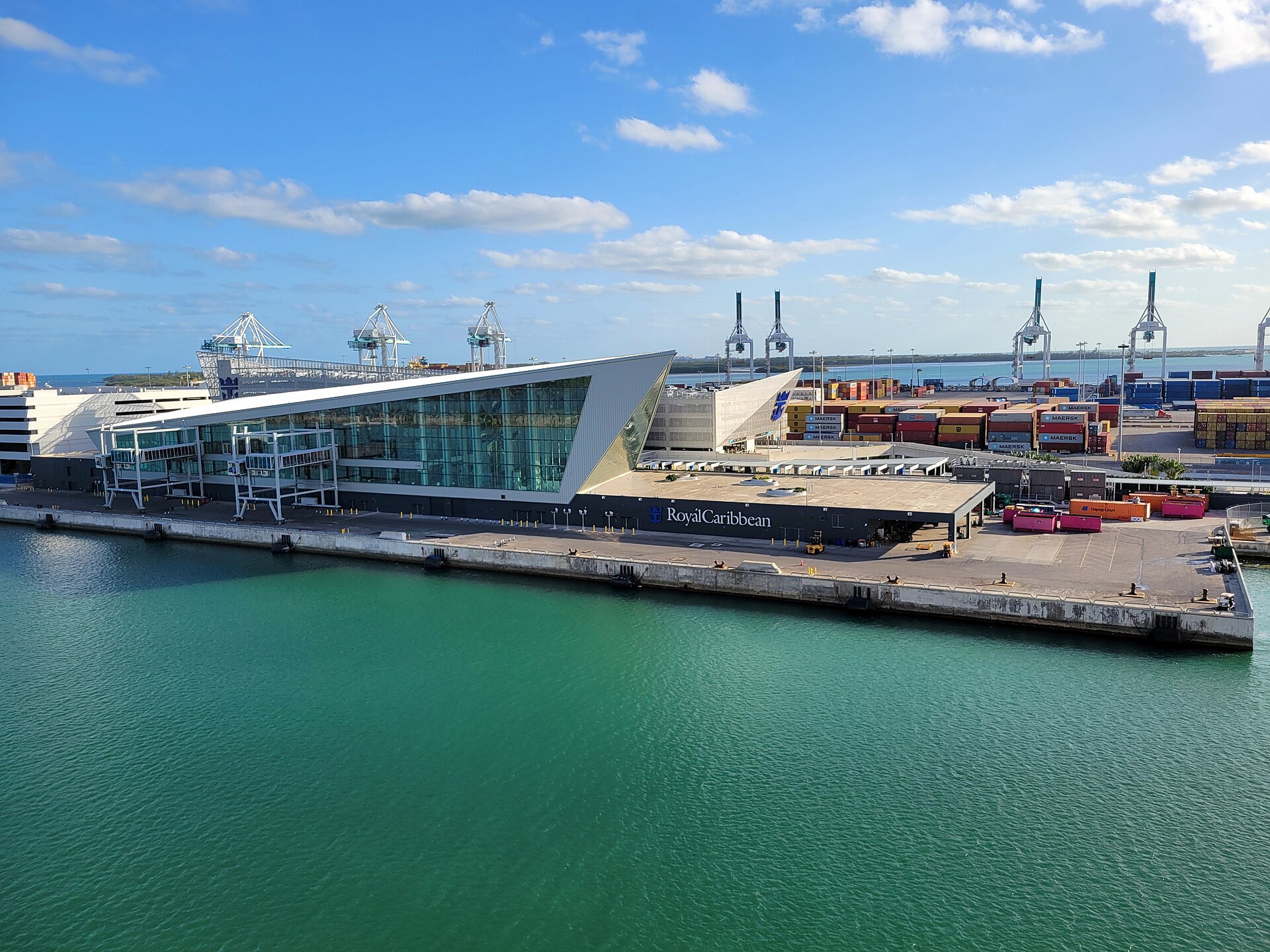 Royal Caribbean's Terminal A at PortMiami, a sharply angled glass building with boarding bridges on the quay and container cranes behind