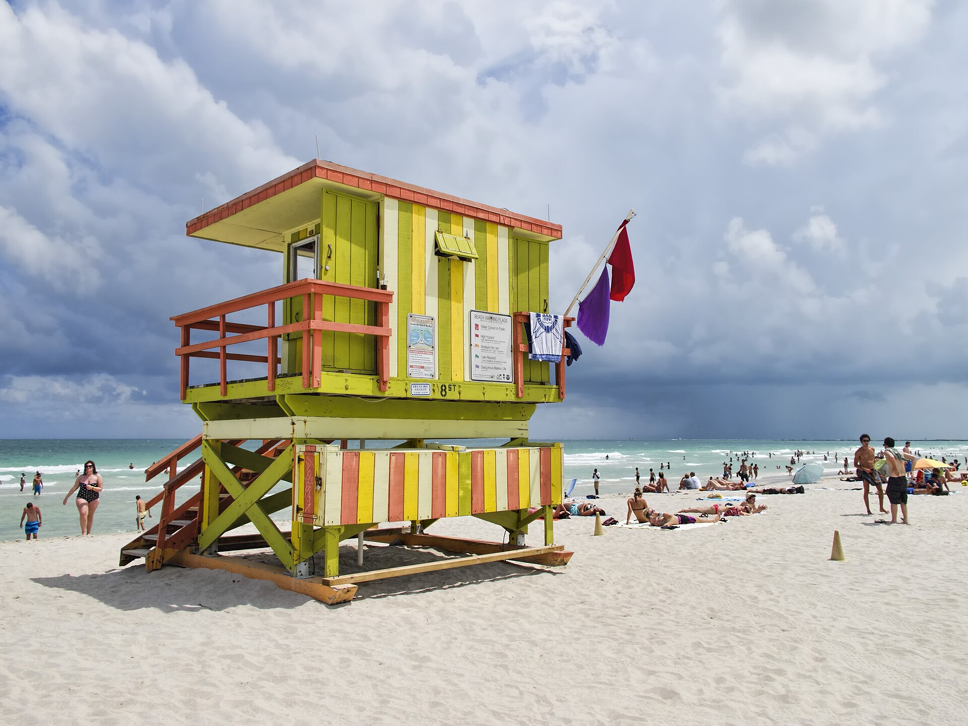 The striped green and orange 8th Street lifeguard stand on South Beach flying red and purple warning flags under a dark sky