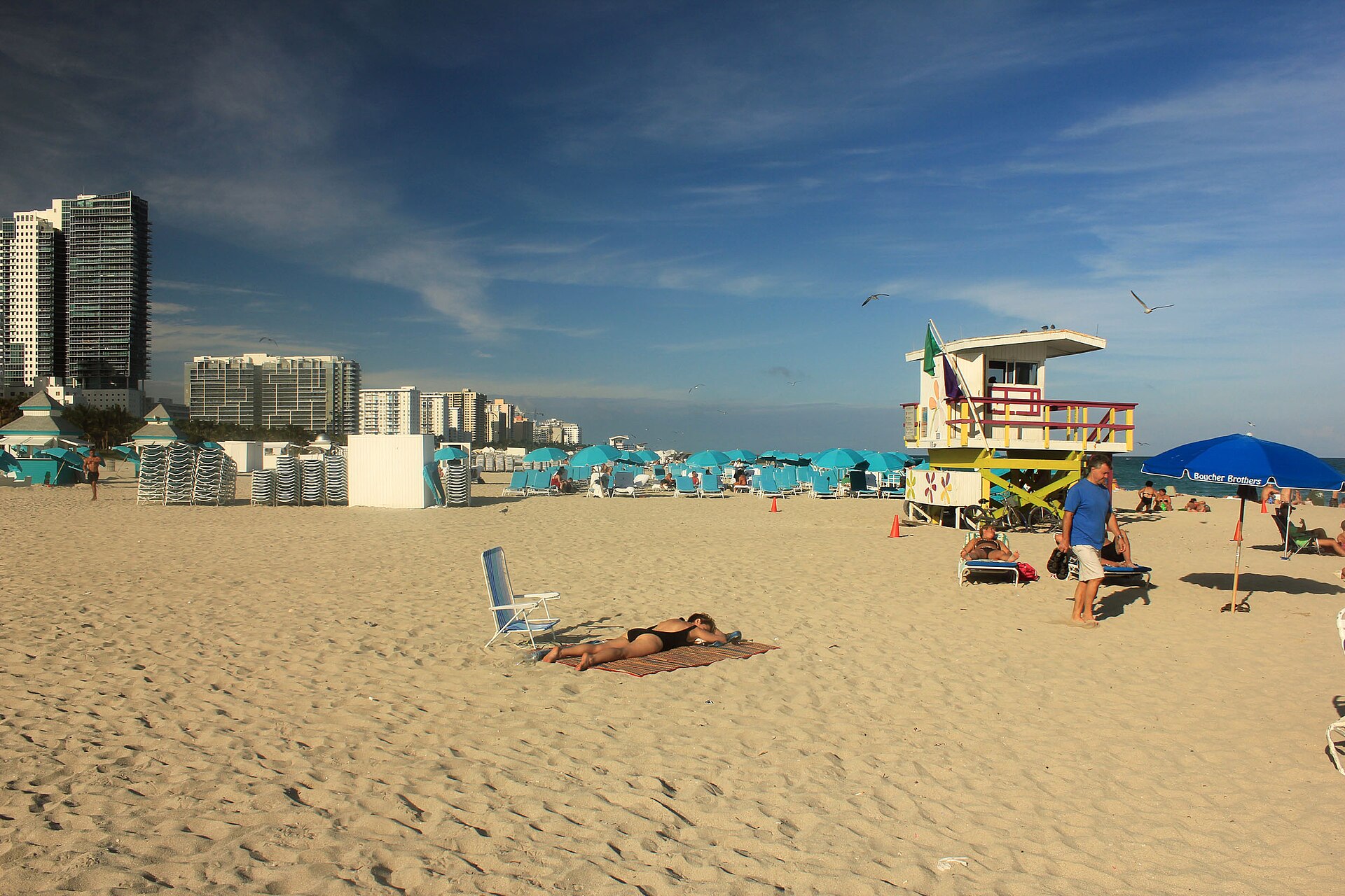 A near-empty stretch of South Beach with a lifeguard tower, rental loungers under teal umbrellas and condo towers in the distance