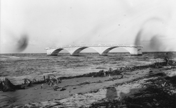 Black-and-white photograph of a low concrete bridge with several arches spanning open water, with seaweed and driftwood on the sand in the foreground