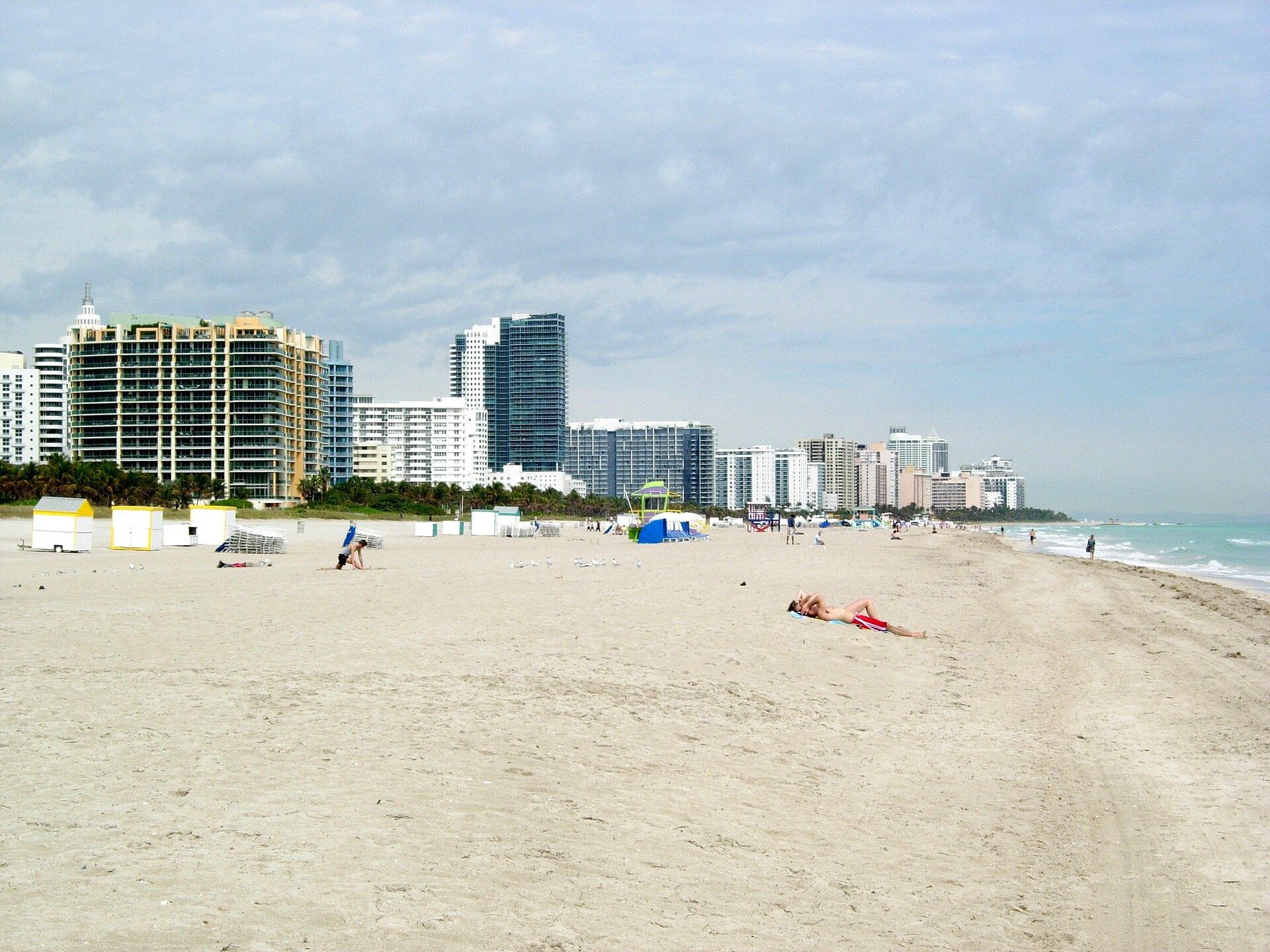 Beachgoers relax on wide South Beach sand in Miami Beach, with ocean surf and beachfront high-rises stretching into the distance.