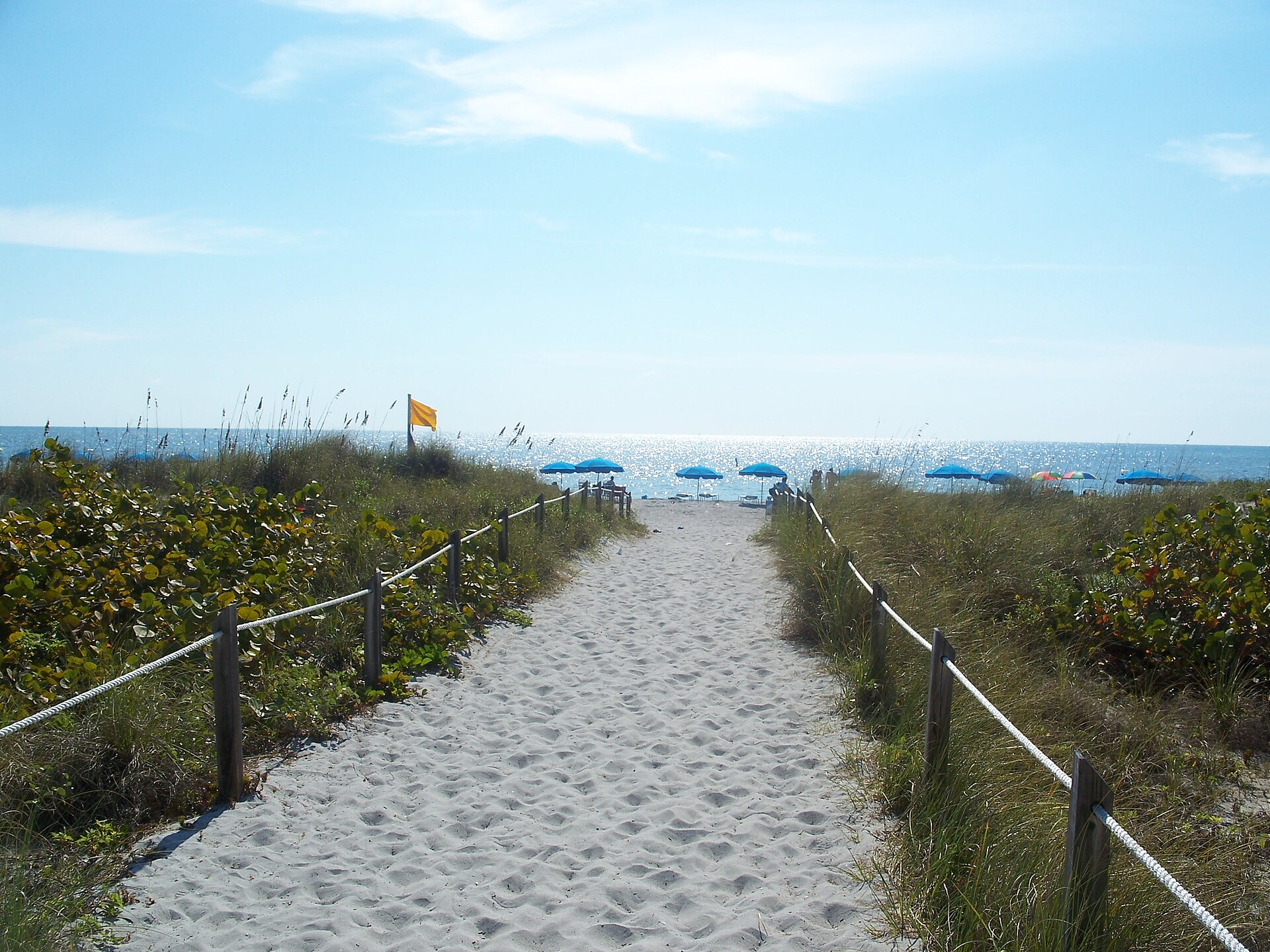 A sandy path between rope fences and sea grape dunes leading to the beach at Bill Baggs, with blue umbrellas, a yellow flag and sparkling sea beyond