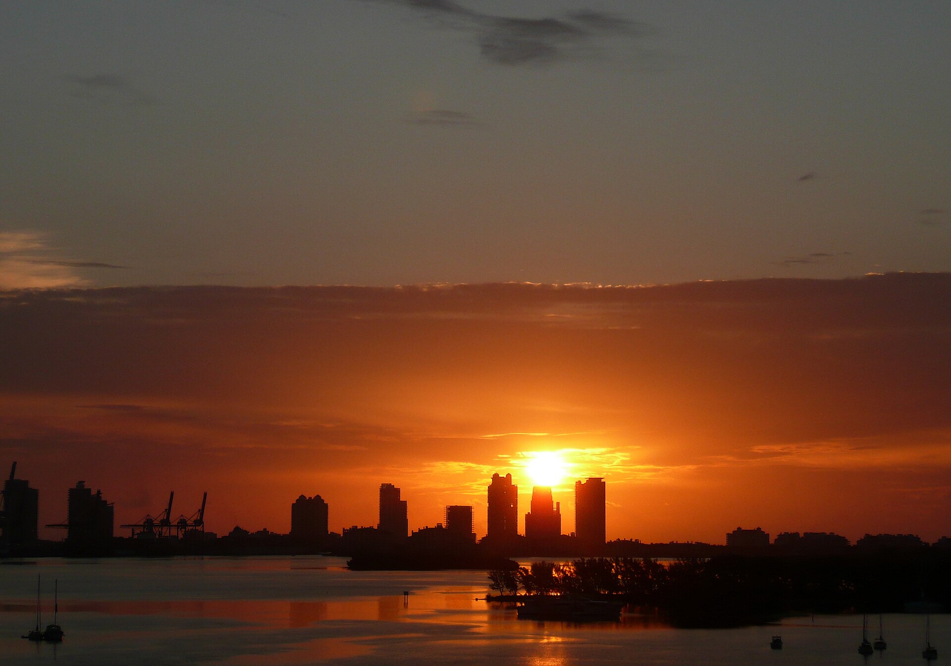 The sun rising behind the towers of South Beach and the PortMiami cranes, silhouetted across the calm water of Biscayne Bay with small boats at anchor
