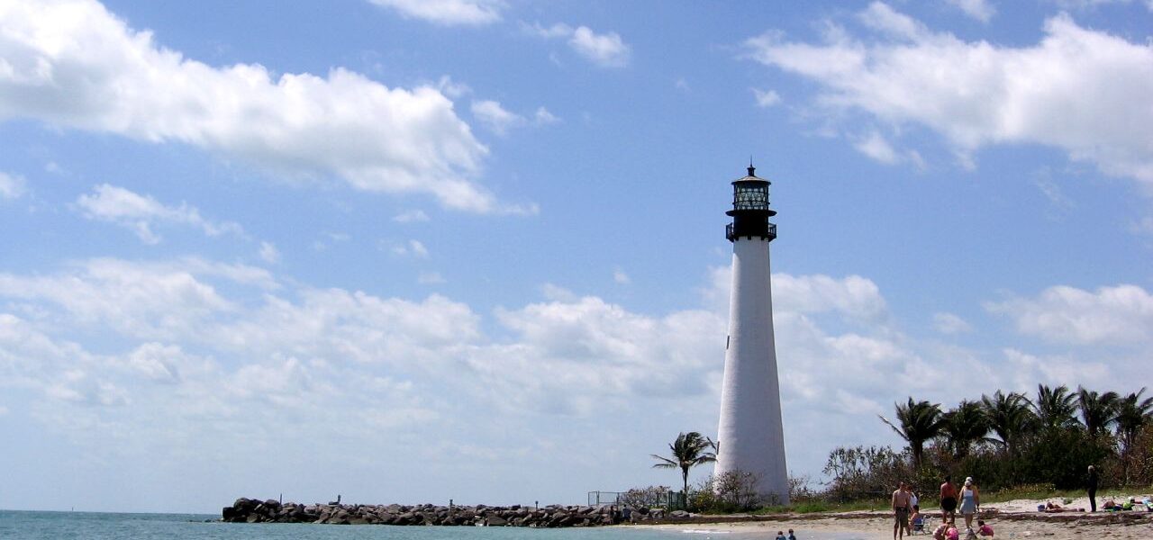 The white Cape Florida Lighthouse rising behind a sandy beach and a rock jetty at Bill Baggs, with a swimmer in clear green water in the foreground