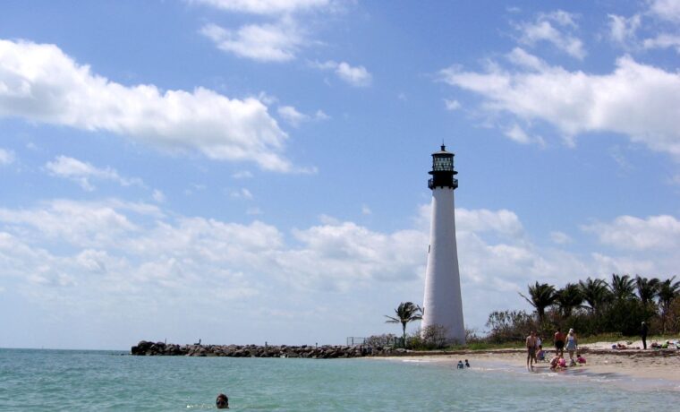 The white Cape Florida Lighthouse rising behind a sandy beach and a rock jetty at Bill Baggs, with a swimmer in clear green water in the foreground