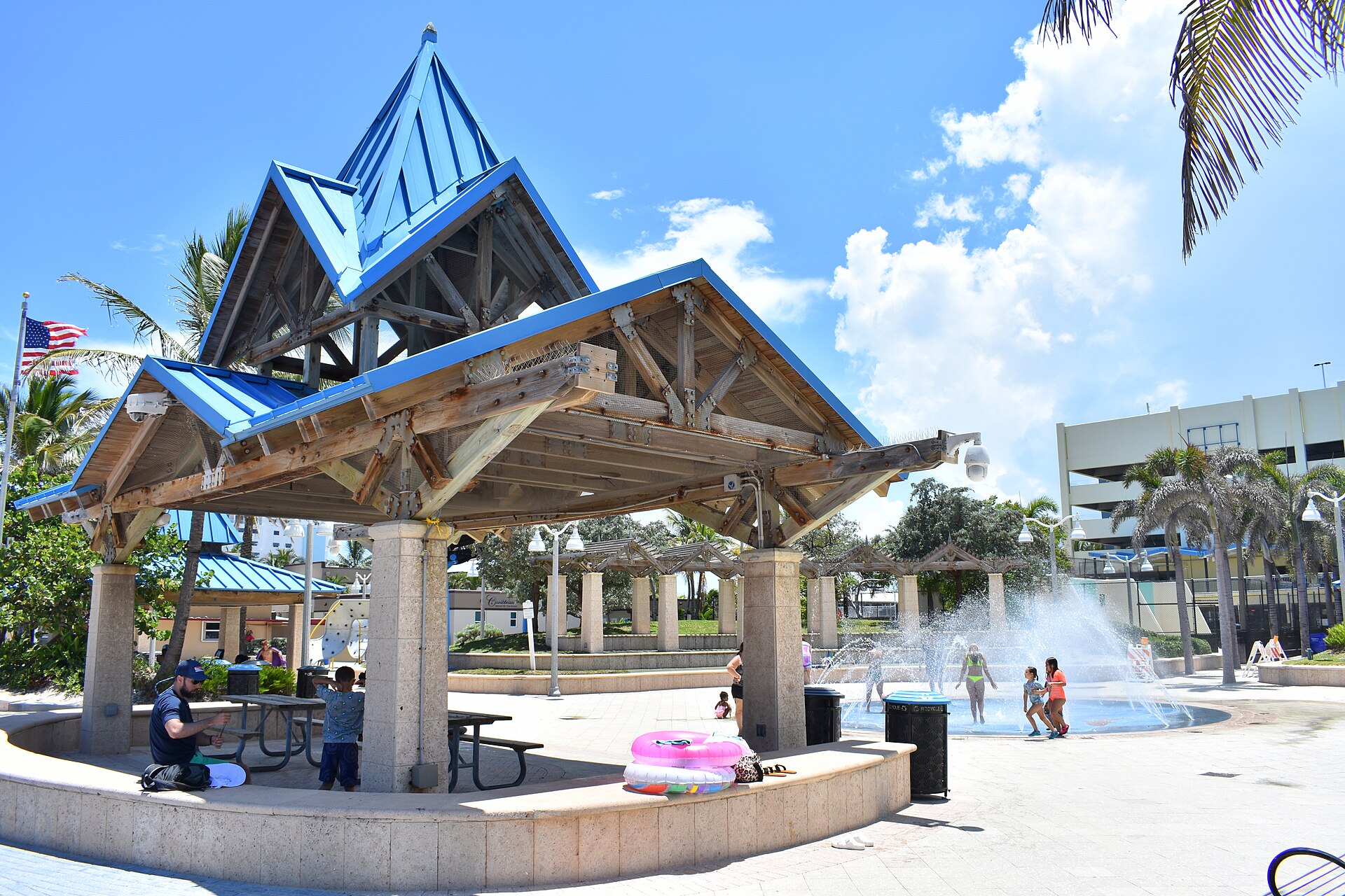 Children playing in the fountains of a splash pad beside a blue-roofed timber picnic pavilion at Charnow Park on Hollywood Beach