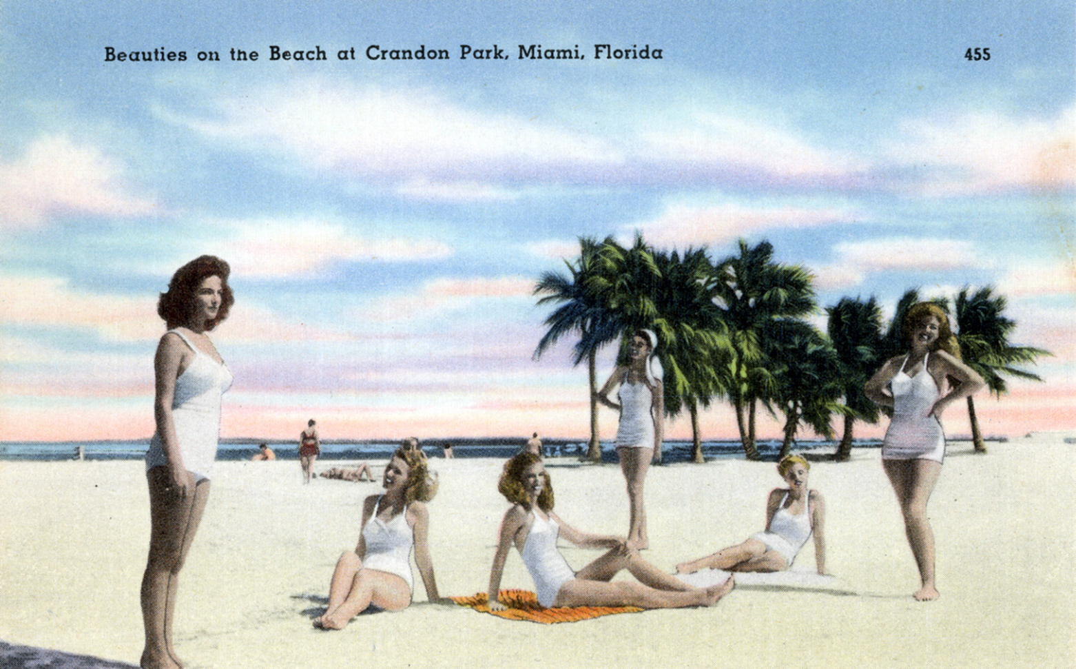 A colorized vintage postcard of five women in white swimsuits posing on the sand at Crandon Park, with palm trees and the sea behind them