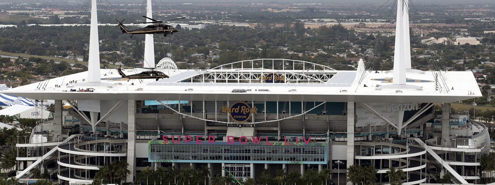 Hard Rock Stadium in Miami Gardens seen from the air, with its white roof, corner masts and helicopters overhead.