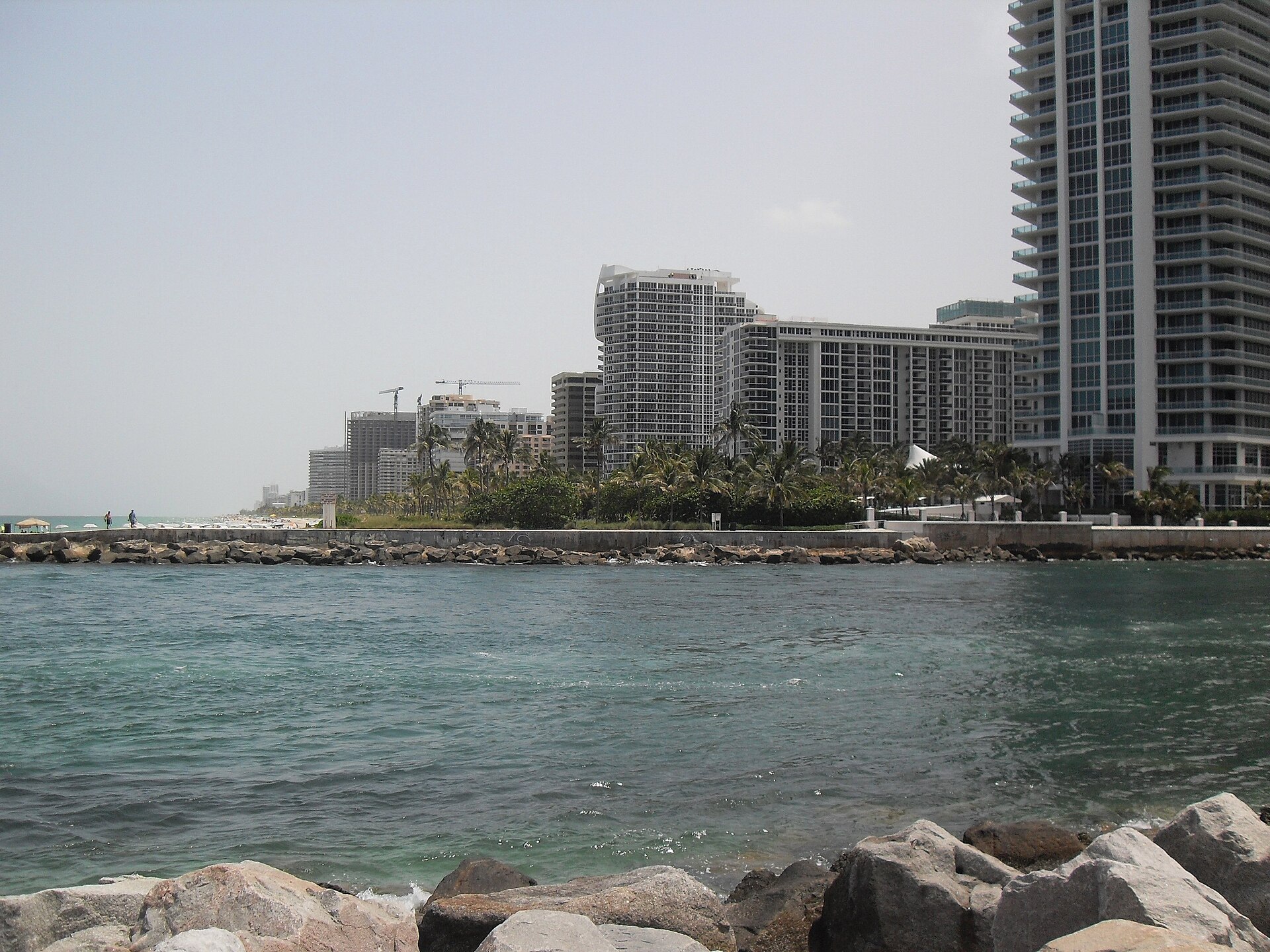 View across Haulover Inlet from the rocks of the jetty to a seawall, palms and the white high-rise towers of Bal Harbour
