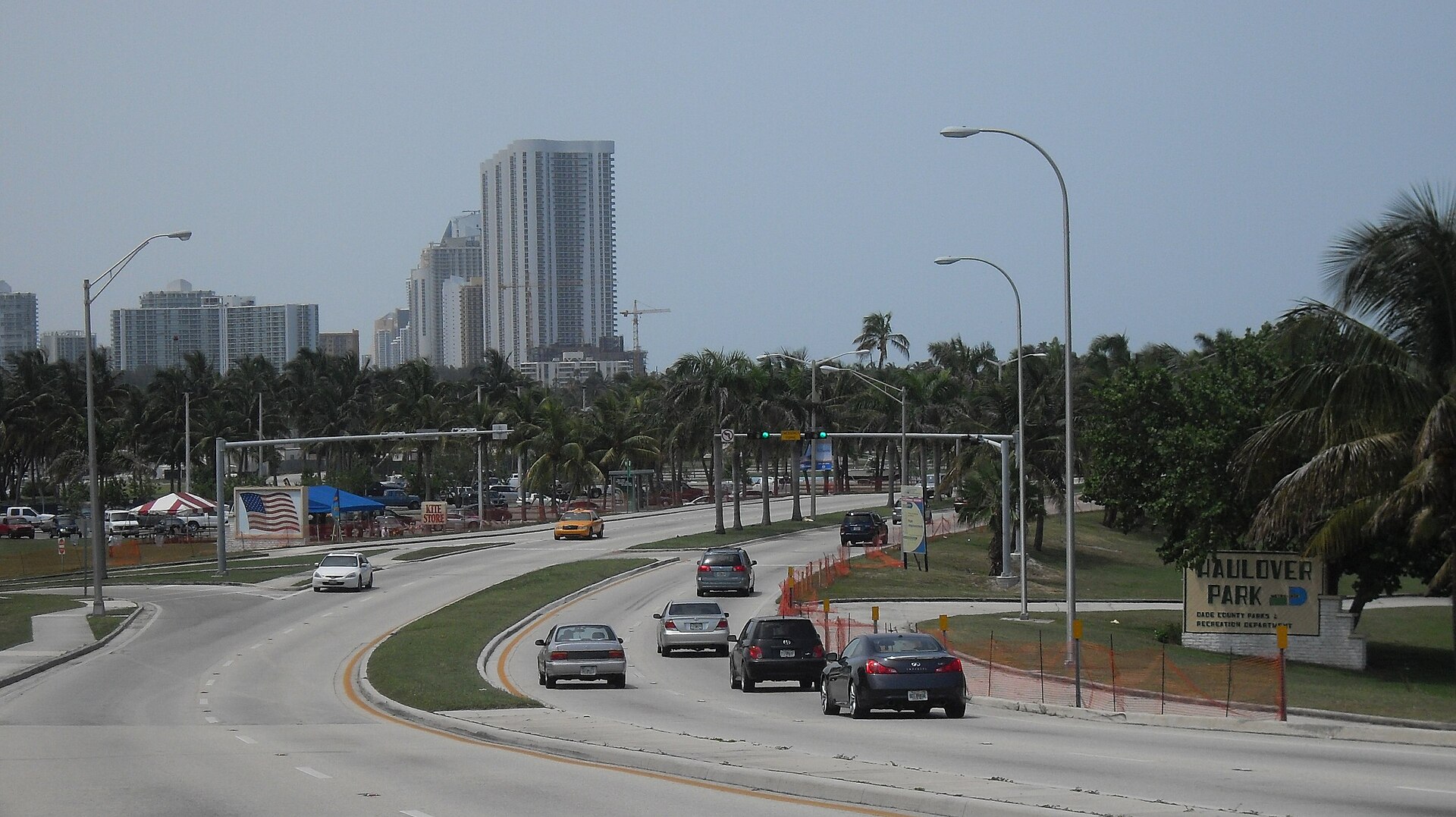 Collins Avenue curving north past the Haulover Park sign, with palms, a kite store stand and parked cars on the left and condo towers in the distance