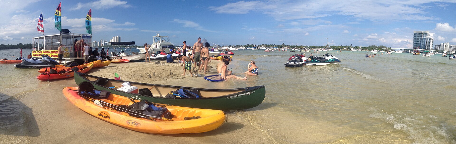 Kayaks, a canoe and jet skis pulled up on a shallow sandbar in Biscayne Bay, with people wading, a floating food vendor and boats anchored all around