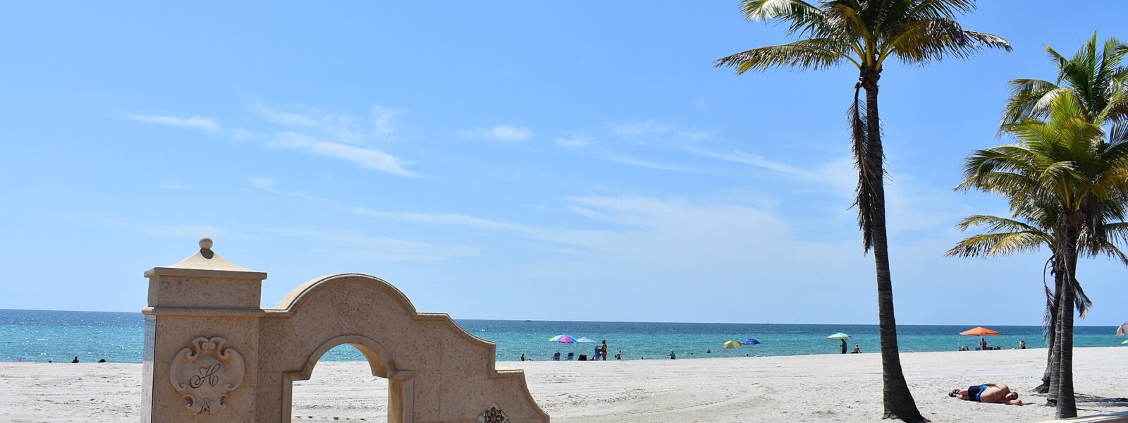 A cream-colored stone gateway with an H crest on the Broadwalk at Hollywood Beach, with coconut palms, wide sand and beach umbrellas by the turquoise sea