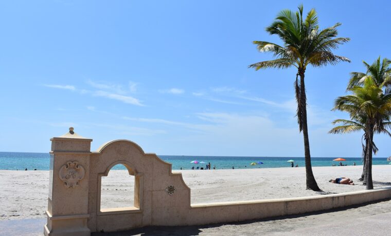 A cream-colored stone gateway with an H crest on the Broadwalk at Hollywood Beach, with coconut palms, wide sand and beach umbrellas by the turquoise sea