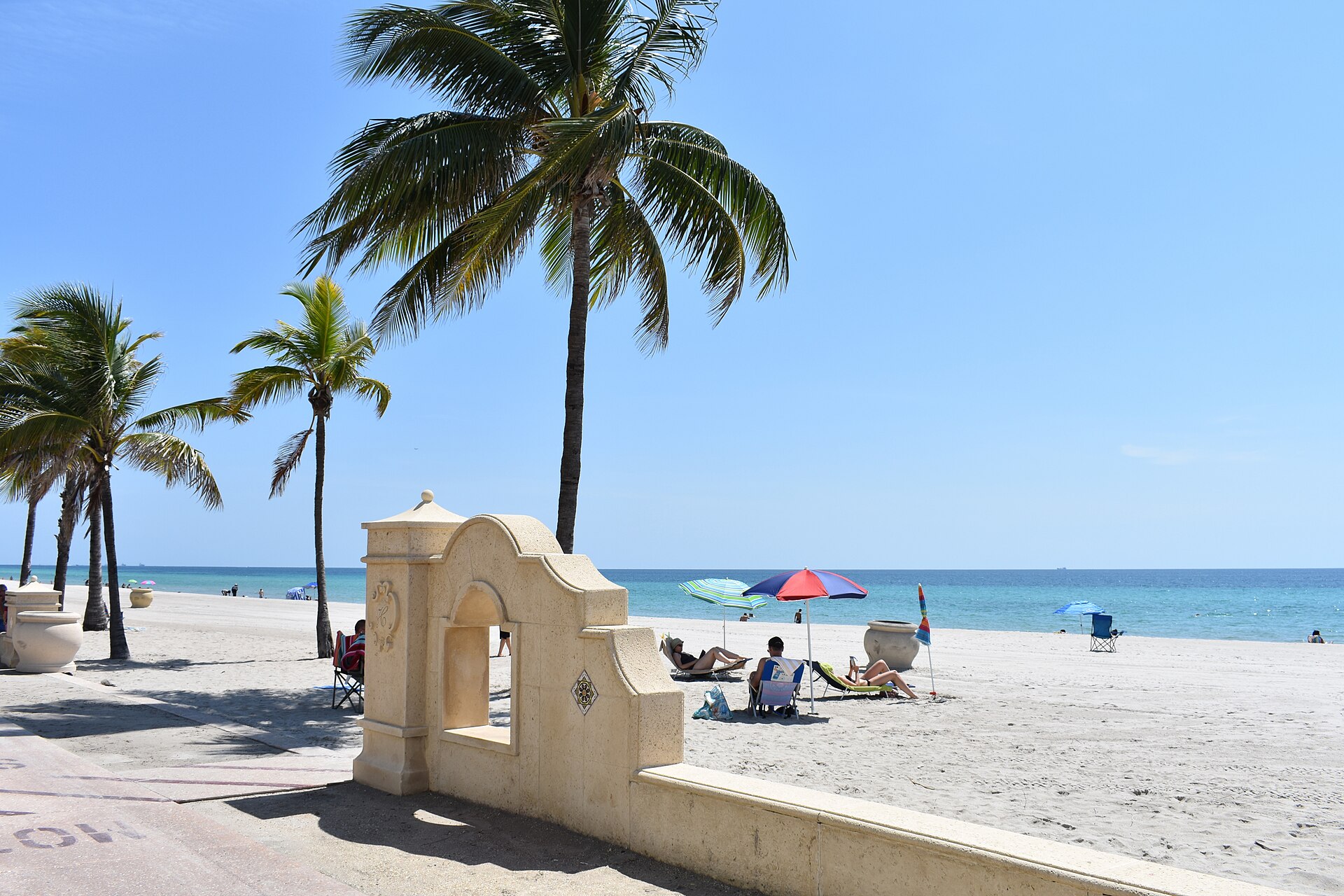 Sunbathers under colorful umbrellas on wide sand beside the Broadwalk at Hollywood Beach, with a cream-colored archway wall decorated with an H crest and tall coconut palms