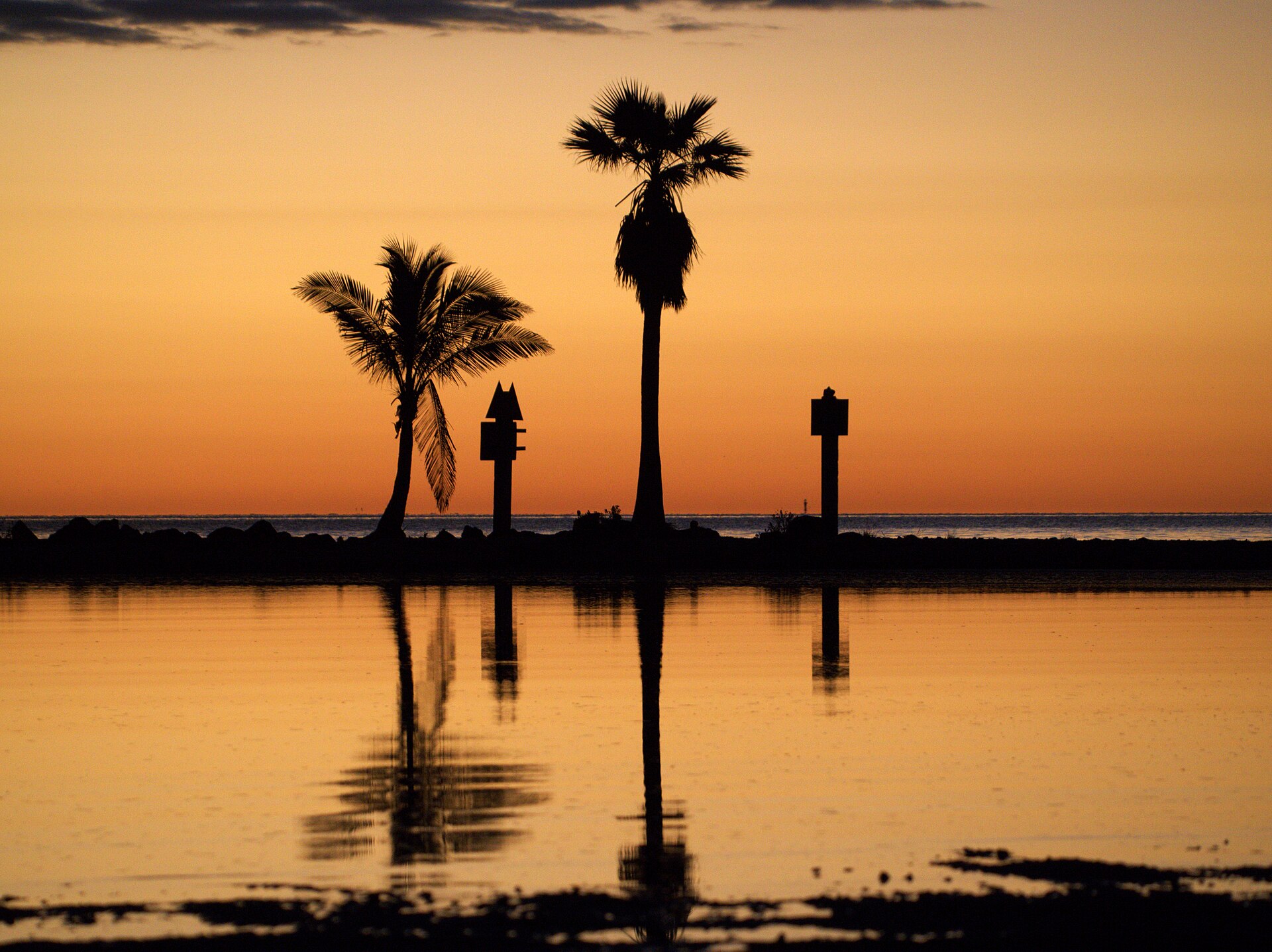 Silhouettes of two palm trees and two channel markers on a low rocky spit, reflected in still water under an orange sky