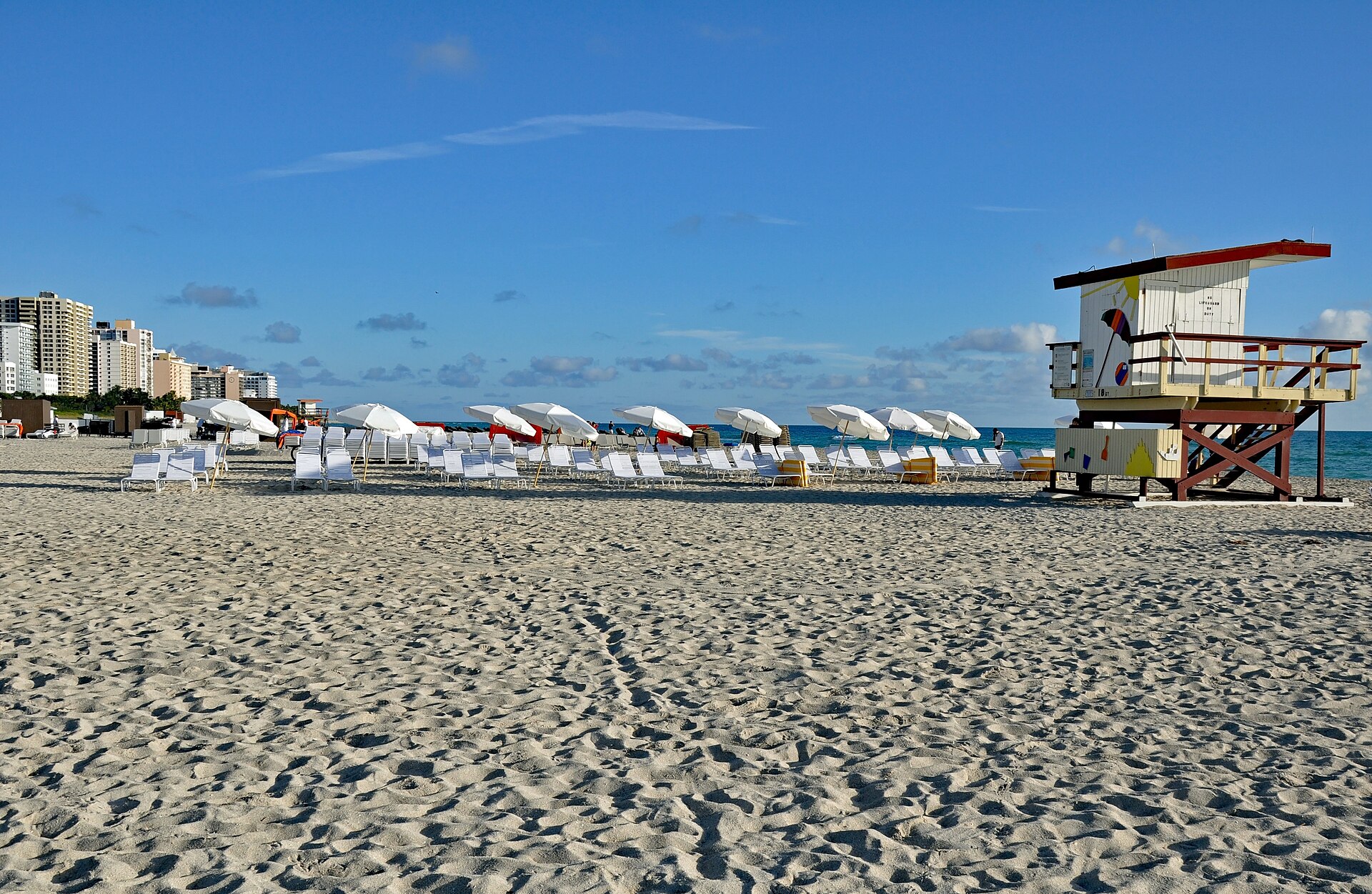 A white lifeguard station with a red roof and painted designs standing on wide sand beside rows of white loungers and umbrellas, with hotels in the distance