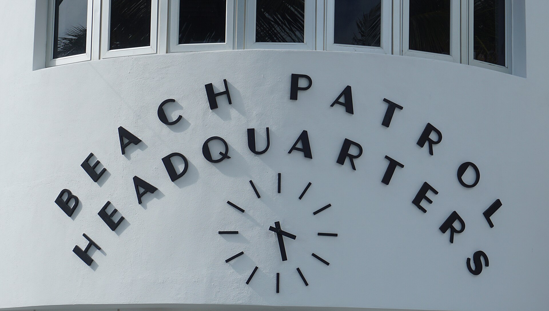 Black Art Deco lettering reading Beach Patrol Headquarters arranged in an arc around a clock face on a curved white wall