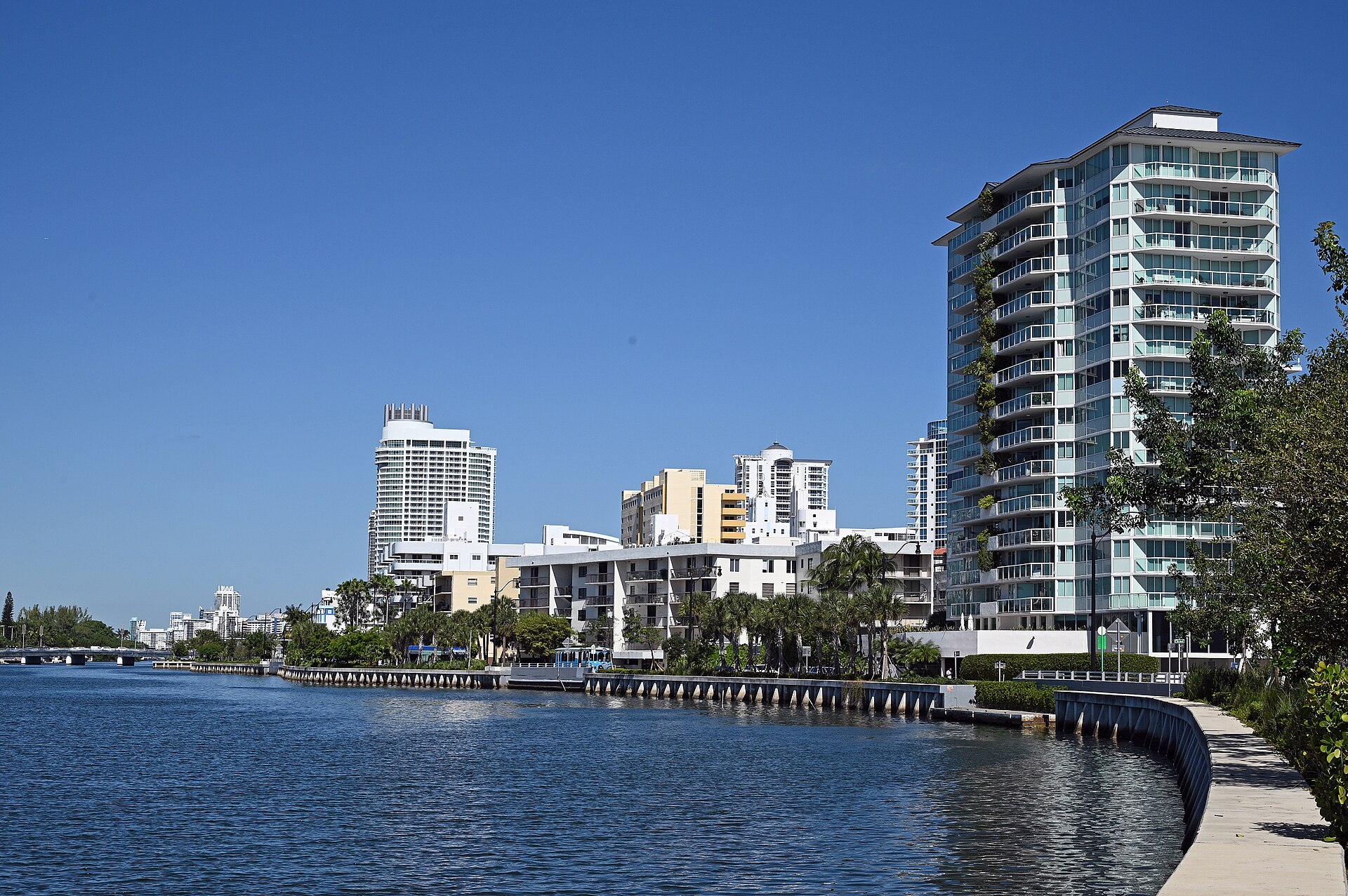 Mid-Beach buildings and palms line Indian Creek in Miami Beach, with a narrow path following the curved seawall.
