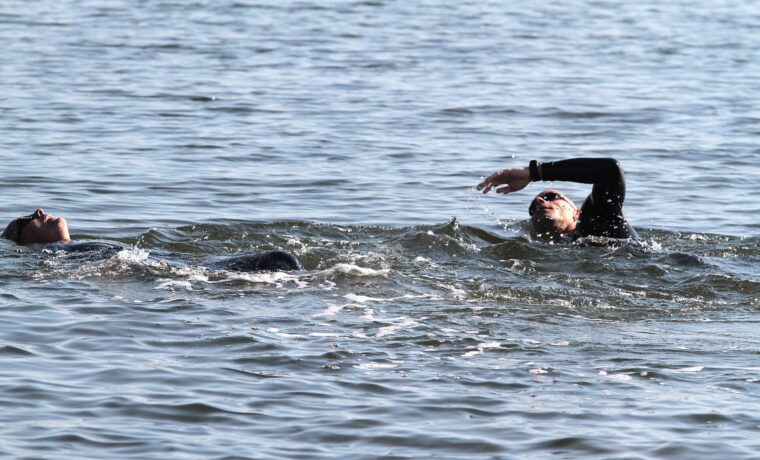 Two open-water swimmers in goggles, one doing freestyle and one floating on his back, in calm sea water